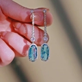 Dettaglio di nappine argentate ed eleganti orecchini con gemma blu ovale e zirconi - Oro Bianco
