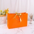 Vibrant orange paper gift bag featuring a golden ribbon bow and sturdy gold rope handles - Orange