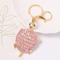Dettaglio di un portachiavi a forma di tartaruga dorata con strass rosa su piatto bianco - Stile Rosa