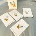 Asymmetric earrings set featuring a white floral heart and brown bear on display cards - Model 01 Woman with Hat