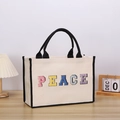 Beige canvas tote bag with black trim and PEACE letter embroidery on wooden surface - PEACE