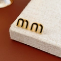 Detail von goldenen M-Ohrringen mit schwarzer Emaille auf beiger strukturierter Fläche - Schwarz