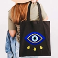 Black canvas tote bag featuring a Greek Evil Eye graffiti print and golden teardrop accents - Design Q-Teardrop Eye-Black