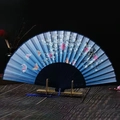 Traditional blue folding fan featuring pink floral and butterfly patterns on a wooden stand - Black Bone Blue Falling Cherry