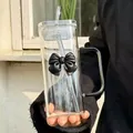 Detail of square glass cup with black bow, handle and straw in a sunlit hand - Black Bow Tie + Straw + Transparent Lid