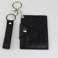 Tarjetero negro con textura tejida, botón de metal y llavero sobre fondo gris minimalista - Negro