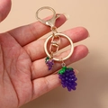Handheld creative purple grape keychain pendant with golden metal ring on beige background - Grapes