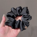 Black satin silk scrunchie with metallic tag shown in hand against a neutral background - Black