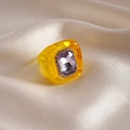 Makro-Detail von gelbem Harzring mit lila Edelstein auf champagnerfarbenem Satinstoff - Gelb und Hellviolett