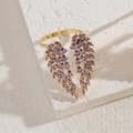 Anello dorato a forma di ala con pietre di zircone viola su sfondo materico - Oro Zirconia Viola