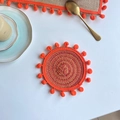 Close-up of hand-woven orange coaster with fringe balls on a white wooden table - 13cm Coaster / Orange