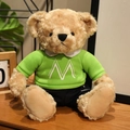 Light brown teddy bear plush in a green 'M' sweater and blue pants on a wooden desk - M Letter Hoodie Set / Green