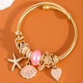 Golden open bangle with starfish shell and heart charms on a warm orange background - Pink