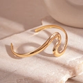 Detail van een golvende gouden manchetarmband met hoogglansafwerking op een stenen blad - Golfvormige open armband / Goud