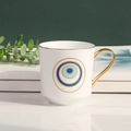 Weiße Evil Eye Keramiktasse mit goldenem Griff und Rand auf minimalistischer Oberfläche - Weiß