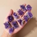 Natürliche violette Amethyst-Cluster und Mineralkristalle in der Hand auf beigem Hintergrund - ca. 10-20g