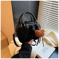Detail shot of a black patent leather crossbody bag with a plush bear charm on white fabric - Black