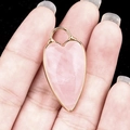 Pink heart quartz pendant with gold plated metal bezel held in hand against black - Rose Quartz