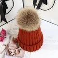 Nahaufnahme der orangefarbenen Strickmütze mit Kunstfell-Bommel neben Geschenktüte - Karamellbraun