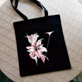 Black canvas tote bag with pink letter V and lily flower print in flat lay view - Letter V / Black