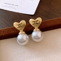 Dettaglio di orecchini a cuore con perle dorate posati su sfondo in legno rustico testurizzato - Orecchino con Perla a Cuore Logo Triomphe (52#) / Oro-Bianco
