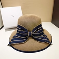 Natural straw sun hat featuring a wide brim and a large navy blue striped bow accent - One Size (Adjustable 55-58.5cm) / Navy Blue Bow / [product Attributes] This Product Is An Exquisite Women's Straw Sun Hat Visually Appearing To Be A Natural/camel Color With A Fine Straw Weave Texture. A Large Dark Blue Bow Adorns One Side Of The Brim With Parallel Gold Stripes Along Its Edges Presenting A Retro Yet Subtly Luxurious Design. [visual Presentation] The Image Is A Well-balanced Indoor Shot. The Background Consists Of A Textured White Gift Box And A Warm Beige Wall Creating Excellent Color Harmony And A Sense Of "breathability." Image Quality Is [high] With Excellent Clarity And Rich Lighting. Aesthetic Appeal Is [high] With An Overall Score Of [8.8]. The Product's Texture Is Fully Enhanced Under Natural Light. [text Content] The Letter [d] Extracted From The Background Gift Box Is [prop_text] And Considered Part Of The Prop; Further Processing Is Not Recommended. [logistics Tags] None [infringement Risk] The Product Itself Has No Logo With A [0.1] Probability Of Infringement. The "d" Logo On The Background Gift Box Is Suspected To Be [dior-background Gift Box-brand Text] Posing A Certain Risk Of Brand Association. 【product Parameters】wide Brim Design Dark Blue Striped Bow Decoration Hard Straw Woven Material.