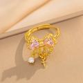 Detail of gold bow ring with pink heart gem and cross pendant on beige background - Gold Pink Ring