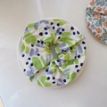 Primer plano detallado de un coletero plisado con estampado floral verde y morado en un plato - Pliegues Irregulares Verde Púrpura sobre Base Blanca
