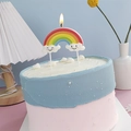 Rainbow cake candle with smiling clouds and flickering flame on pink surface - Rainbow Cloud Candle