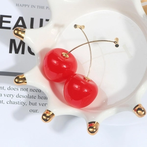 Detailed shot of vibrant red cherry earrings with gold hooks on a white porcelain tray