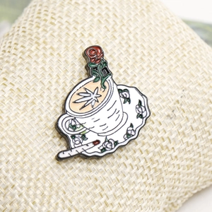 Macro detail of enamel coffee cup brooch with rose and cannabis leaf on woven fabric