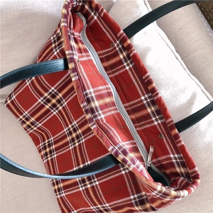 Macro view of red plaid tote bag featuring zipper closure and black leather straps