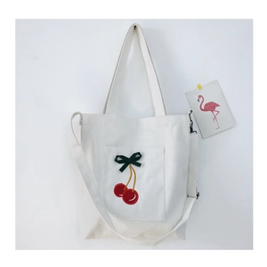 Close-up of a white canvas tote bag featuring an embroidered red cherry and green bow