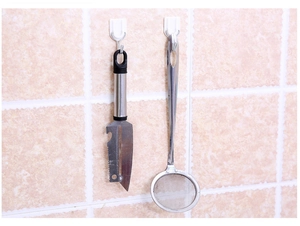 Close-up of white plastic adhesive wall hooks holding a peeler and kitchen utensil on tiles