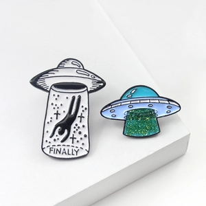 Detail view of alien UFO enamel pins with FINALLY text and glitter light beam on white stand