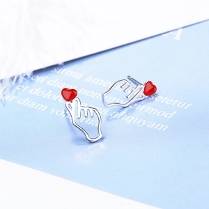 Minimalist silver hand gesture stud earrings with red heart accent on blue background