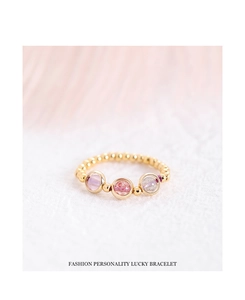 Makroaufnahme eines Goldperlenarmbands mit rosa und lila Kristallen auf weicher Feder