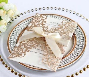 Detail of glitter laser-cut gold wedding invitation with beige ribbon on decorative plate