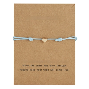 Close-up of light blue woven cord bracelet with gold heart charm on kraft card