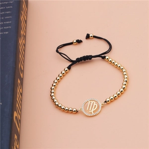 Close-up of Virgo zodiac bracelet with gold beads and black braided rope on peach background