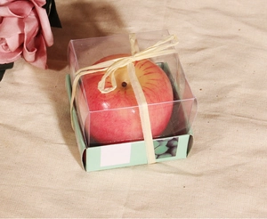 Detalle de vela de manzana roja en caja PVC transparente con lazo de rafia natural