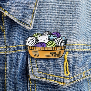 Cute white cat in colorful yarn basket enamel pin pinned on a blue denim jacket pocket