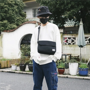 Street style view of a black canvas messenger bag worn by a model against a garden wall