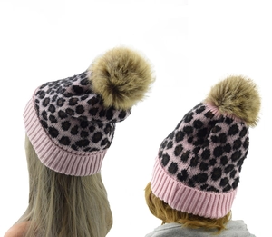 Close-up of leopard print knit beanies featuring pink ribbed cuffs and large faux fur pom poms
