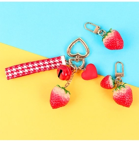 Detail of strawberry keychain featuring a red bell, heart clasp, and checkered ribbon