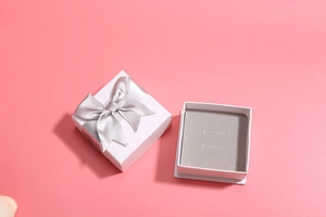 Two white square jewelry boxes with silver ribbon bows and grey sponge lining on pink base