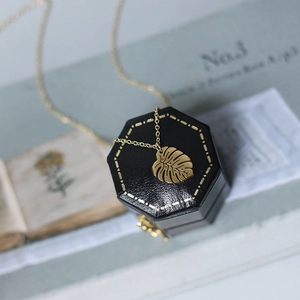 Golden monstera leaf necklace displayed on a black jewelry box with vintage book background