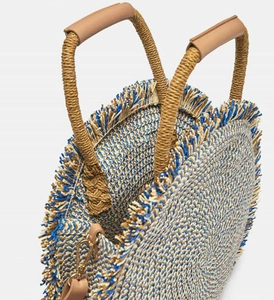Close-up of a round woven straw bag with blue accents and premium brown leather handles