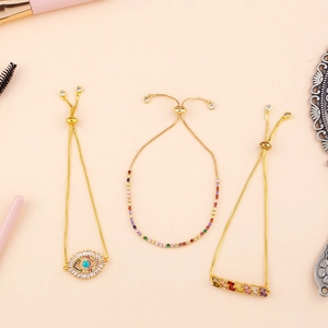 Adjustable gold bracelets with evil eye and rainbow cubic zirconia charms on peach background