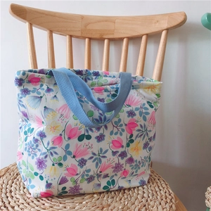 Florale Canvas-Tragetasche mit blauen Griffen und zartem Druck auf hellem Stuhl